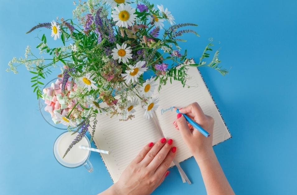 A woman's hand journaling with a bouquet of flowers to unlock abundance and improve her money mindset.